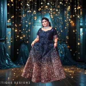 May include: A woman wearing a long, sequined gown with a blue to rose gold ombre effect. The gown has a fitted bodice and a flowing skirt. The woman is standing in front of a backdrop of blue curtains and twinkling lights.