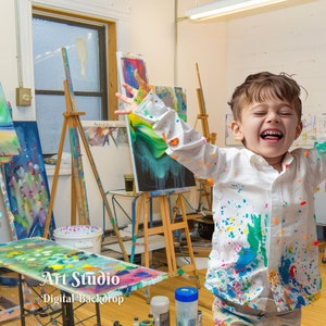 May include: A young boy wearing a white shirt covered in colorful paint splatters stands in an art studio with several easels and paintings. The boy is smiling and has his arms raised in the air. The text "Art Studio - Digital Backdrop" is visible in the lower left corner of the image.