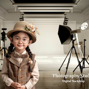 Puede incluir: Una persona joven con un sombrero marrón con un adorno floral, un chaleco a cuadros y gafas posa en un estudio fotográfico. El estudio tiene equipo de iluminación profesional con reflectores negros y fondos blancos.