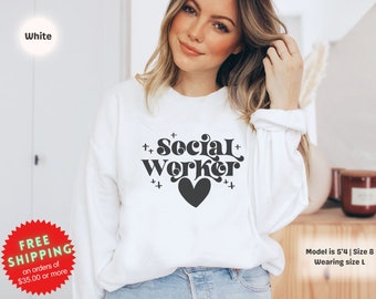 May include: White sweatshirt with the text "++Social+Worker++" in black with a black heart in the center. The model is wearing a size large.
