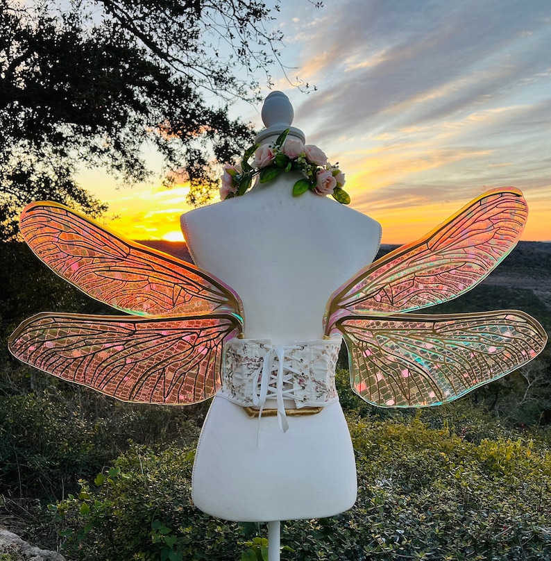 K&ouml;nnte beinhalten: Ein Paar irisierende Libellenfl&uuml;gel mit einem goldenen Drahtrahmen, befestigt an einem wei&szlig;en Korsett mit einem Spitzenmuster. Die Fl&uuml;gel werden auf einem wei&szlig;en Mannequin vor einem Sonnenuntergangs-Hintergrund pr&auml;sentiert.