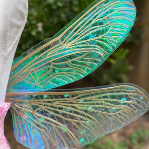 K&ouml;nnte beinhalten: Ein Paar regenbogenfarbene gr&uuml;ne Feenfl&uuml;gel mit goldenen Adern und Glitzerakzenten. Die Fl&uuml;gel sind aus einem durchscheinenden Material gefertigt und haben einen zarten, &auml;therischen Look.