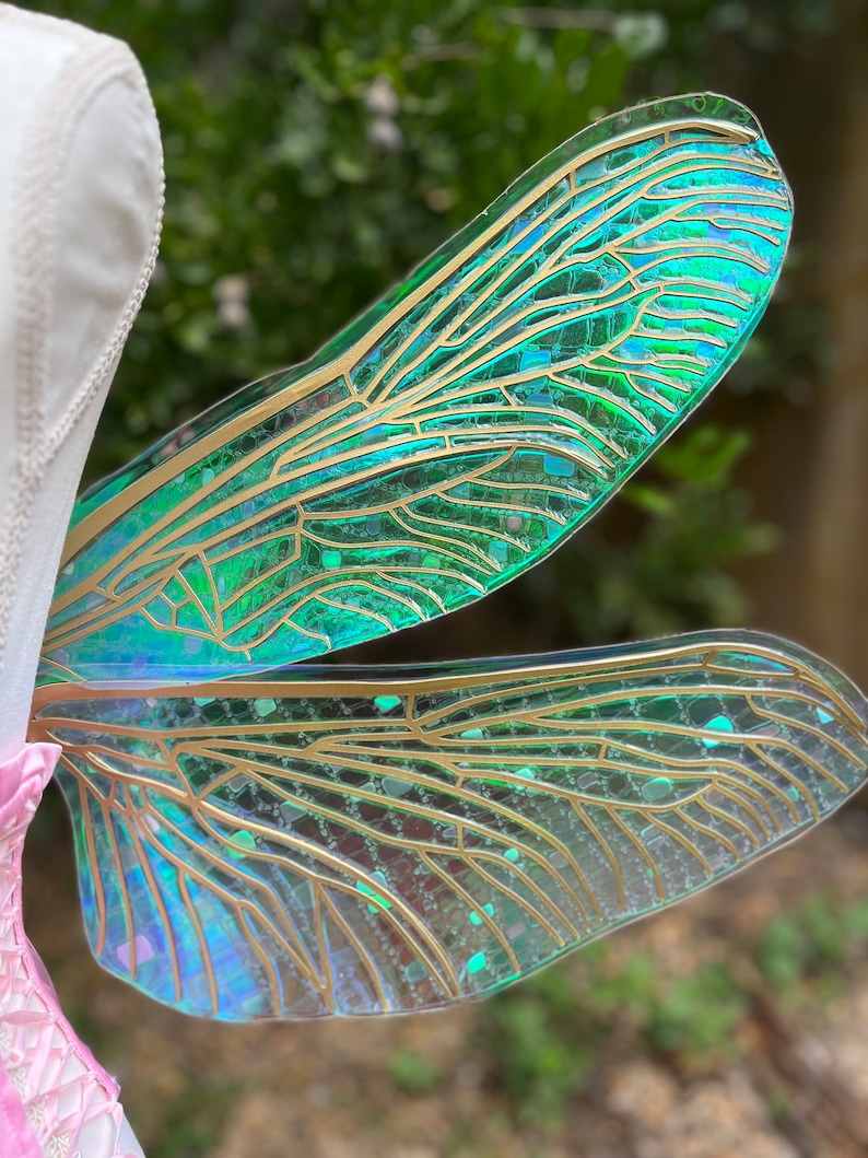 K&ouml;nnte beinhalten: Ein Paar regenbogenfarbene gr&uuml;ne Feenfl&uuml;gel mit goldenen Adern und Glitzerakzenten. Die Fl&uuml;gel sind aus einem durchscheinenden Material gefertigt und haben einen zarten, &auml;therischen Look.