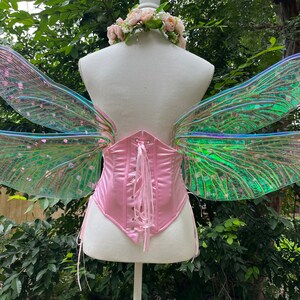 K&ouml;nnte beinhalten: Ein Paar regenbogenfarbene gr&uuml;ne und rosa Libellen-Feenfl&uuml;gel mit einem rosa Satin-Korsett. Die Fl&uuml;gel sind an einer wei&szlig;en Puppe befestigt.