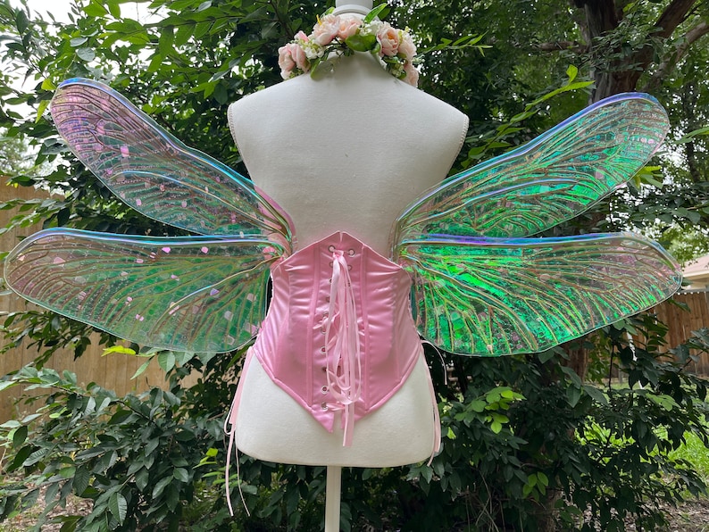 K&ouml;nnte beinhalten: Ein Paar regenbogenfarbene gr&uuml;ne und rosa Libellen-Feenfl&uuml;gel mit einem rosa Satin-Korsett. Die Fl&uuml;gel sind an einer wei&szlig;en Puppe befestigt.