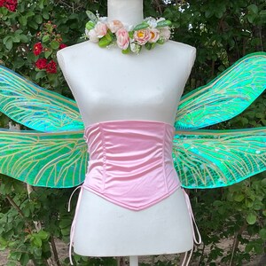 K&ouml;nnte beinhalten: Ein Paar regenbogenfarbene gr&uuml;ne Feenfl&uuml;gel mit goldenen Adern, befestigt an einem rosa Satin-Korsett. Die Fl&uuml;gel befinden sich auf einer wei&szlig;en Schaufensterpuppe mit einer Blumenkrone.