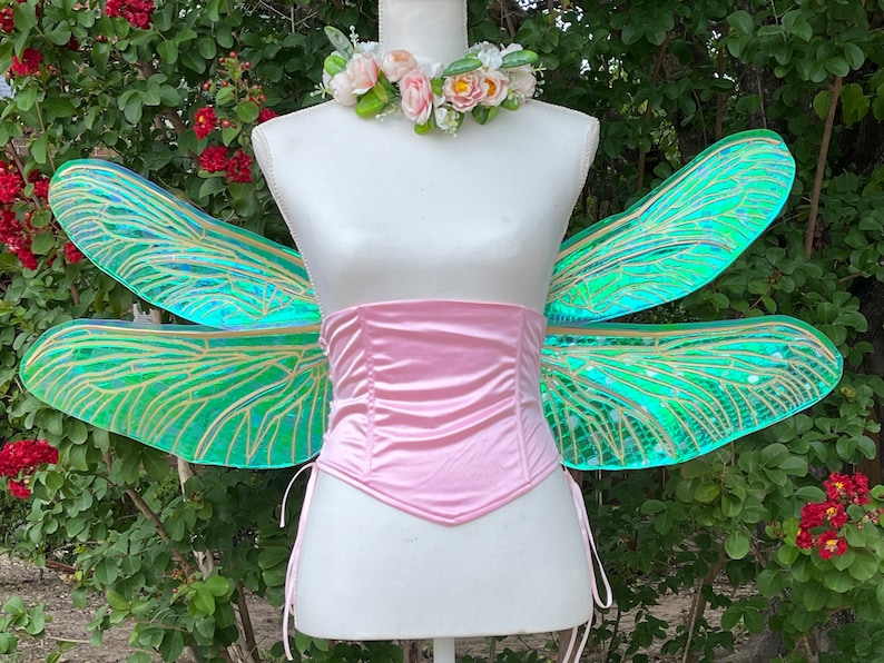 K&ouml;nnte beinhalten: Ein Paar regenbogenfarbene gr&uuml;ne Feenfl&uuml;gel mit goldenen Adern, befestigt an einem rosa Satin-Korsett. Die Fl&uuml;gel befinden sich auf einer wei&szlig;en Schaufensterpuppe mit einer Blumenkrone.