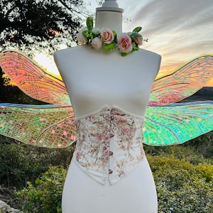 K&ouml;nnte beinhalten: Eine wei&szlig;e Schaufensterpuppe tr&auml;gt ein florales Korsett und schillernde Libellenfl&uuml;gel. Die Fl&uuml;gel sind hellgr&uuml;n, rosa und blau. Die Schaufensterpuppe tr&auml;gt eine Blumenkrone mit rosa und wei&szlig;en Blumen.