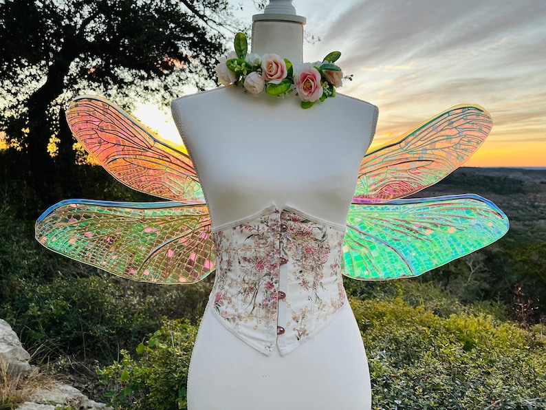 K&ouml;nnte beinhalten: Eine wei&szlig;e Schaufensterpuppe tr&auml;gt ein florales Korsett und schillernde Libellenfl&uuml;gel. Die Fl&uuml;gel sind hellgr&uuml;n, rosa und blau. Die Schaufensterpuppe tr&auml;gt eine Blumenkrone mit rosa und wei&szlig;en Blumen.