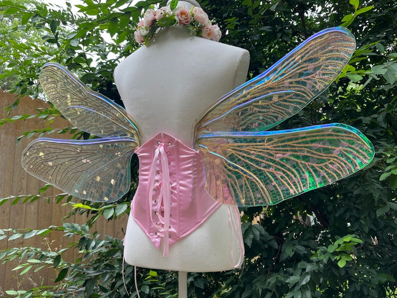 K&ouml;nnte beinhalten: Ein Paar regenbogenfarbene Feenfl&uuml;gel mit einem rosa Satin-Korsett. Die Fl&uuml;gel sind aus klarem Kunststoff mit einem zarten Muster und haben eine schimmernde Oberfl&auml;che.