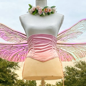 K&ouml;nnte beinhalten: Rosa schimmernde Libellenfl&uuml;gel mit einem rosa Satin-Korsett. Die Fl&uuml;gel sind an einer wei&szlig;en Schaufensterpuppe befestigt.