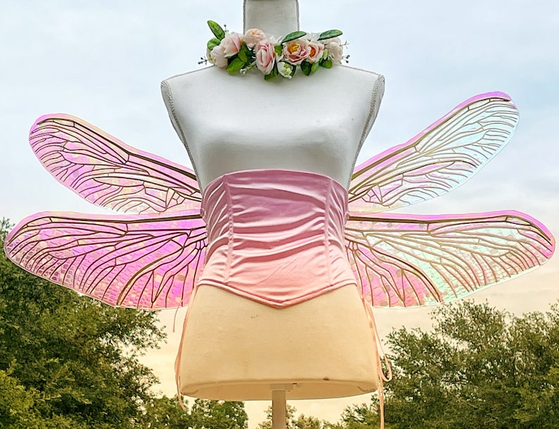 K&ouml;nnte beinhalten: Rosa schimmernde Libellenfl&uuml;gel mit einem rosa Satin-Korsett. Die Fl&uuml;gel sind an einer wei&szlig;en Schaufensterpuppe befestigt.