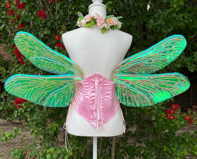 K&ouml;nnte beinhalten: Ein Paar regenbogenfarbene gr&uuml;ne Libellenfl&uuml;gel mit einem rosa Satin-Korsett. Die Fl&uuml;gel sind aus einem durchscheinenden Material mit einem zarten Adernmuster gefertigt.