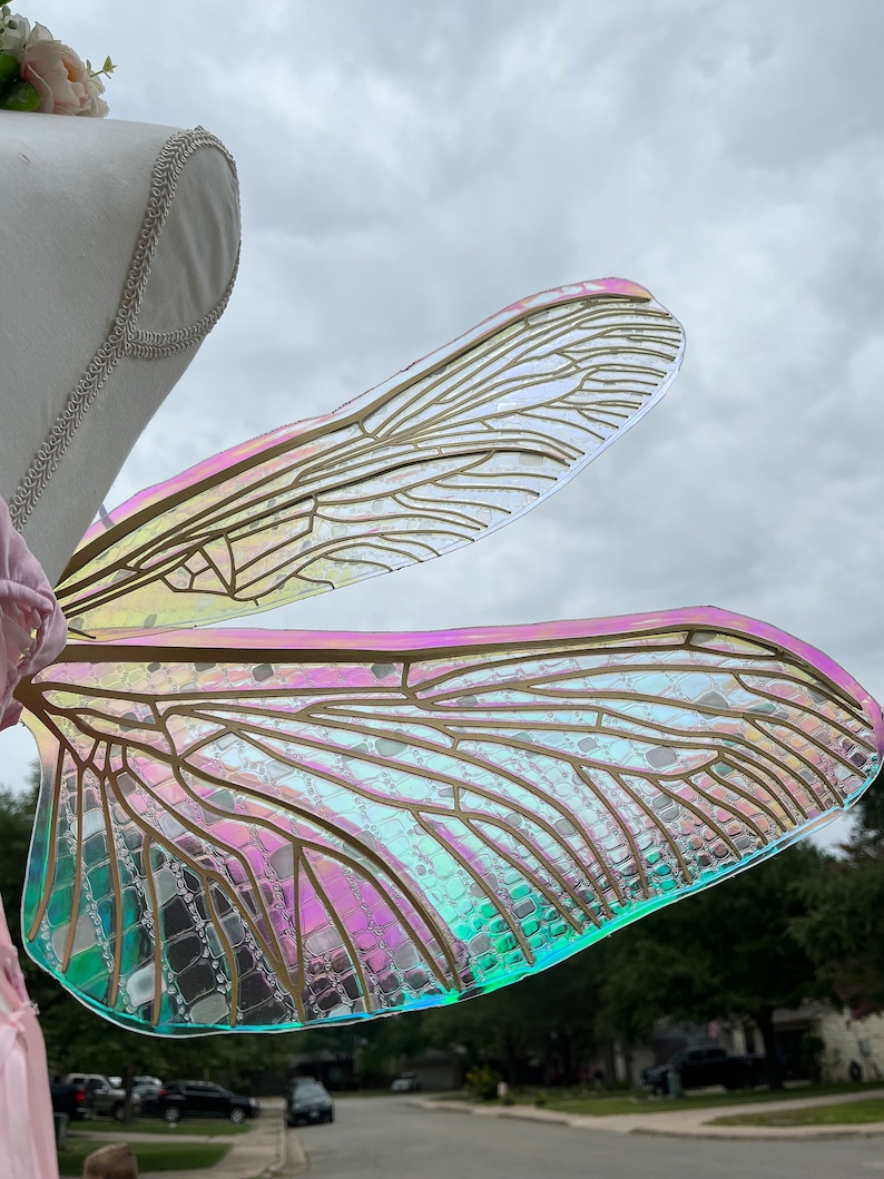 K&ouml;nnte beinhalten: Ein Paar regenbogenfarbene Feenfl&uuml;gel mit einem goldenen Drahtrahmen. Die Fl&uuml;gel bestehen aus einem klaren Kunststoffmaterial mit holografischem Finish. Die Fl&uuml;gel sind an einem wei&szlig;en Mannequin befestigt.