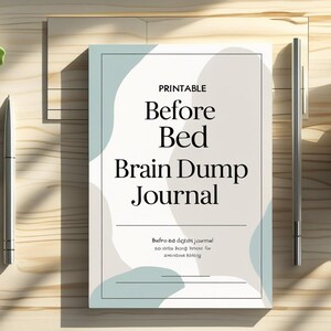 Puede incluir: Un "Diario de descarga cerebral antes de dormir" con una cubierta azul claro y blanca, un bolígrafo, una taza de té y suculentas sobre una superficie de madera. El título del diario está en texto negro. Es un artículo imprimible.