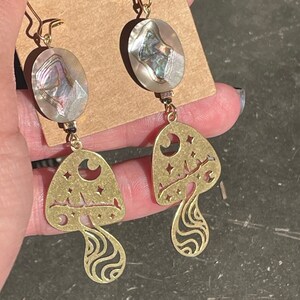 May include: A pair of gold-tone earrings featuring a mushroom design with a crescent moon and stars. The top of each earring is a round iridescent abalone shell.