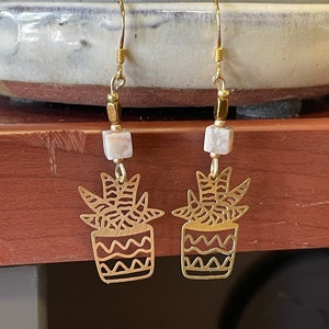 May include: A pair of gold-tone earrings featuring a cutout design of a potted succulent plant. Each earring has a white and gray square bead hanging from the top.