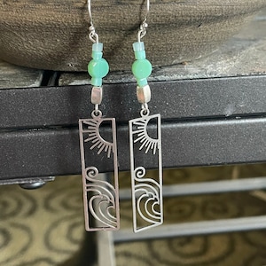 May include: Silver dangle earrings with a sun and wave design. The earrings have a light green bead at the top.