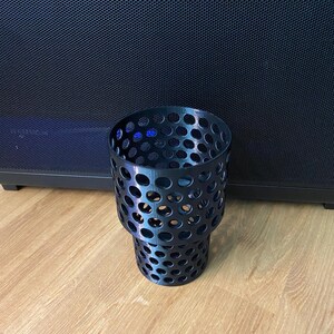 Puede incluir: Un portavasos negro, cil&iacute;ndrico, con un dise&ntilde;o perforado. El portavasos est&aacute; hecho de un material oscuro y presenta numerosos agujeros circulares. Se encuentra sobre una superficie de madera, con un fondo oscuro.
