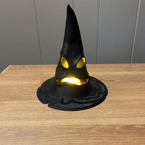 May include: A black, pointed witch hat with a wide brim, illuminated from within by a warm, yellow light. The hat has a textured surface and is sitting on a wooden table. The hat's eye-shaped cutouts glow.
