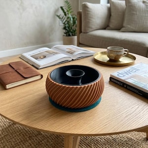 May include: A round wooden coffee table with a brown spiral-patterned bowl, a leather-bound notebook, an open book, a gold tray with a cup, and a magazine. The bowl sits on a teal coaster. A sofa and plant are in the background.