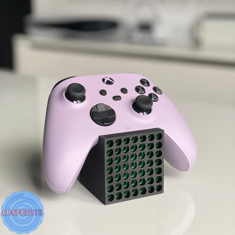 May include: A light purple Xbox controller rests on a black stand with a grid of green-filled holes. The controller has black joysticks and buttons. The stand is on a white surface.