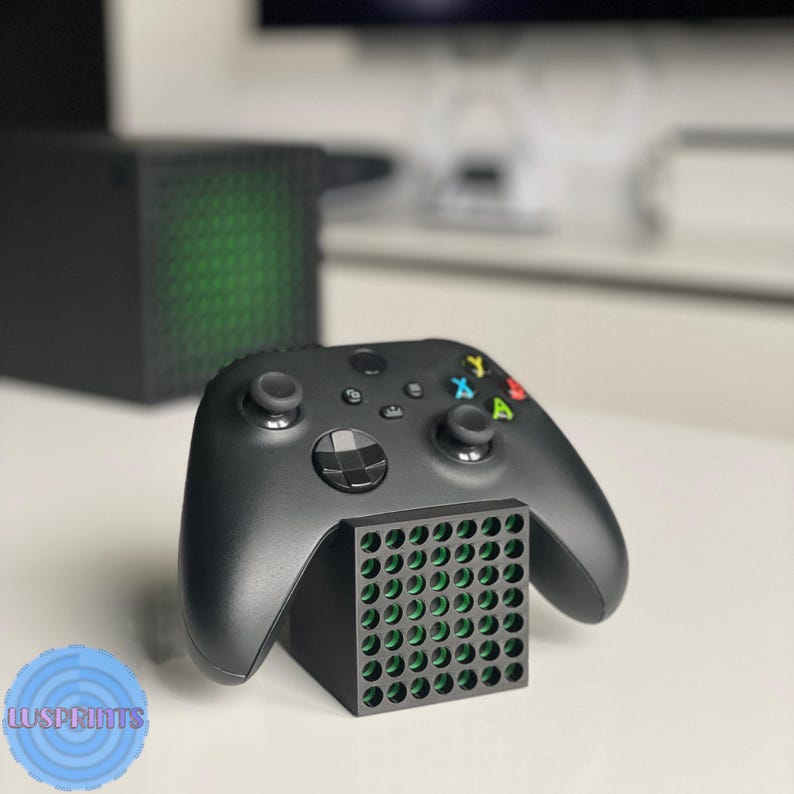 May include: Black Xbox controller resting on a black stand with a grid pattern. The controller has colored buttons and analog sticks. An Xbox Series X console is visible in the background.