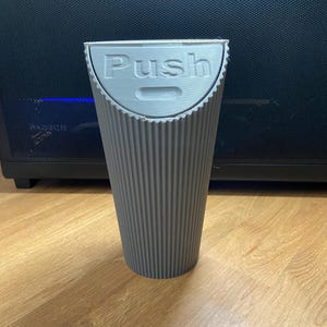 May include: A gray, ribbed, cylindrical container with a hinged lid. The word "Push" is embossed on the lid. The container is made of a light gray material and sits on a wooden surface.