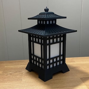 May include: Black pagoda-style lantern with a tiered roof and square, open sides. The lantern has a decorative finial on top and sits on a wooden surface. The lantern's design is reminiscent of traditional Asian architecture.