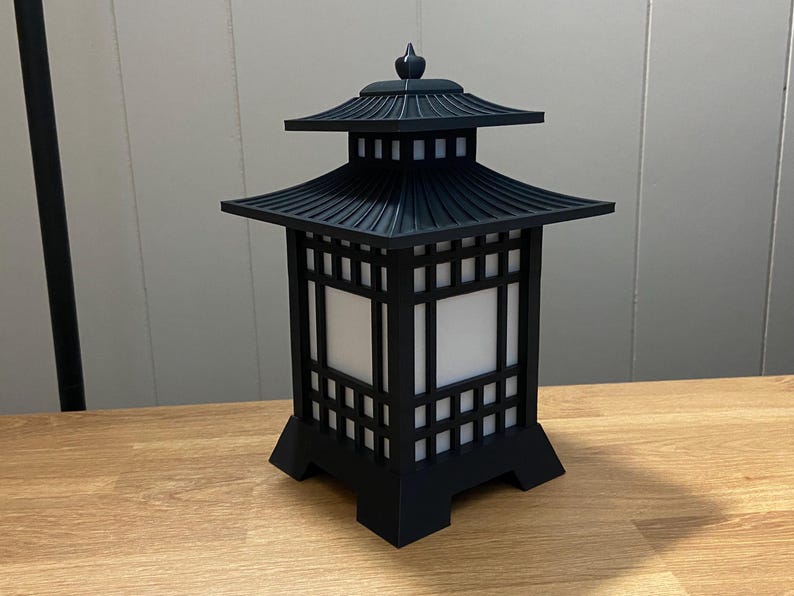 May include: Black pagoda-style lantern with a tiered roof and square, open sides. The lantern has a decorative finial on top and sits on a wooden surface. The lantern's design is reminiscent of traditional Asian architecture.