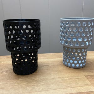 Puede incluir: Dos recipientes cil&iacute;ndricos con un dise&ntilde;o perforado. Uno es negro, el otro gris. Los recipientes est&aacute;n apilados, con la secci&oacute;n superior ligeramente m&aacute;s peque&ntilde;a que la inferior. Est&aacute;n sobre una superficie de madera.