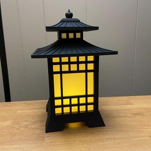 May include: Black Japanese-style lantern with a pagoda-style roof and a square, grid-like design. The lantern has a warm, yellow glow emanating from within. It is made of a dark material and sits on a wooden surface.