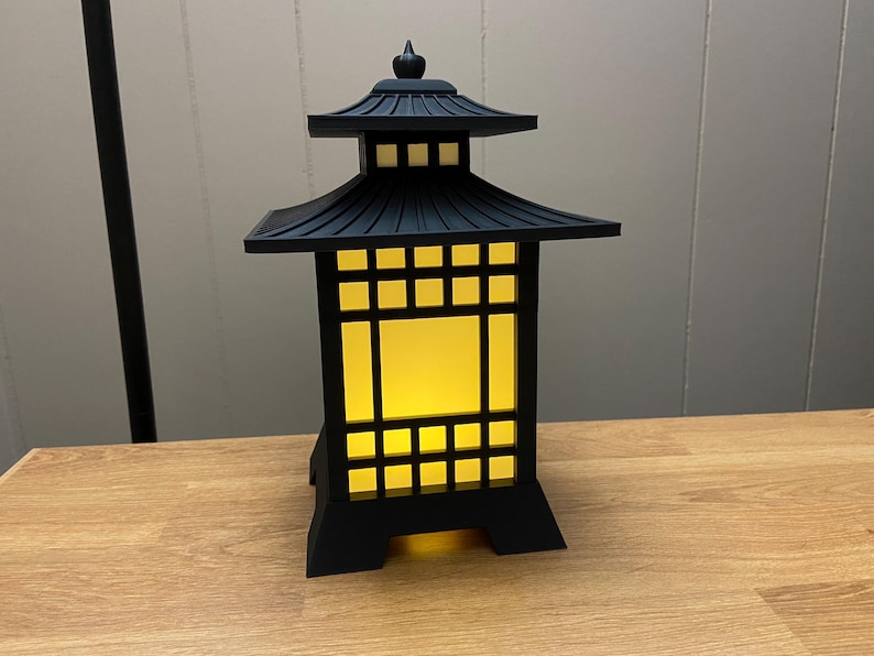 May include: Black Japanese-style lantern with a pagoda-style roof and a square, grid-like design. The lantern has a warm, yellow glow emanating from within. It is made of a dark material and sits on a wooden surface.