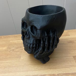 Peut inclure: Un pot de fleurs noir en forme de cr&acirc;ne avec un int&eacute;rieur creux. Le pot pr&eacute;sente des traits faciaux d&eacute;taill&eacute;s et une surface textur&eacute;e. La base est soutenue par une main stylis&eacute;e.