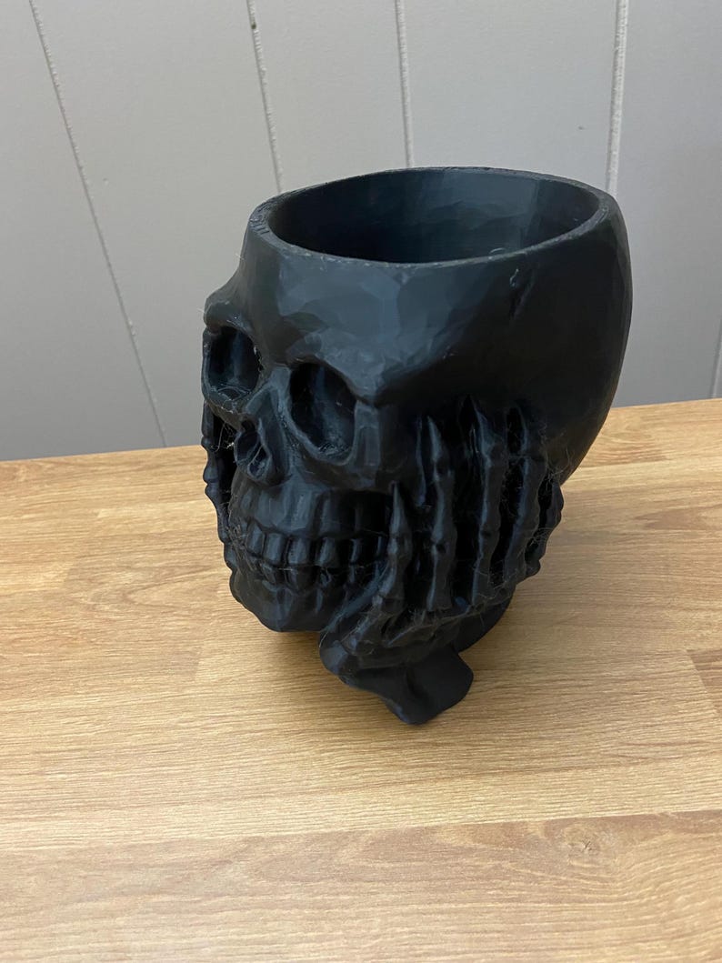 Peut inclure: Un pot de fleurs noir en forme de cr&acirc;ne avec un int&eacute;rieur creux. Le pot pr&eacute;sente des traits faciaux d&eacute;taill&eacute;s et une surface textur&eacute;e. La base est soutenue par une main stylis&eacute;e.