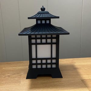 May include: A black, Japanese-style lantern with a pagoda-style roof and a square, grid-patterned body. The lantern has a white, illuminated interior and sits on a wooden surface. The lantern is a decorative lighting fixture.
