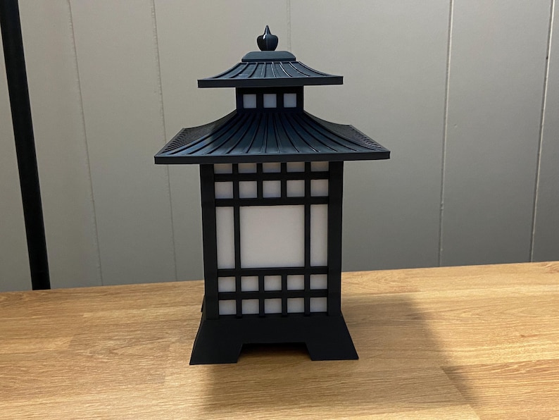 May include: A black, Japanese-style lantern with a pagoda-style roof and a square, grid-patterned body. The lantern has a white, illuminated interior and sits on a wooden surface. The lantern is a decorative lighting fixture.