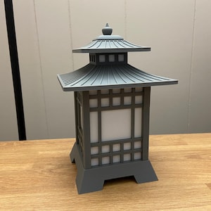 May include: A grey pagoda-style lantern with a tiered roof and a square base. The lantern has a grid-like design on the sides with a white, illuminated panel in the centre. The lantern is made of a solid material and sits on a wooden surface.