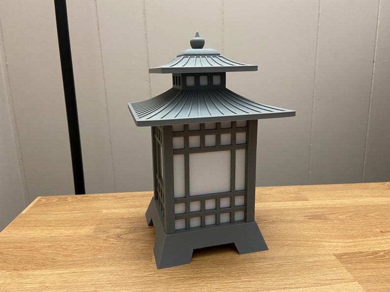 May include: A grey pagoda-style lantern with a tiered roof and a square base. The lantern has a grid-like design on the sides with a white, illuminated panel in the centre. The lantern is made of a solid material and sits on a wooden surface.