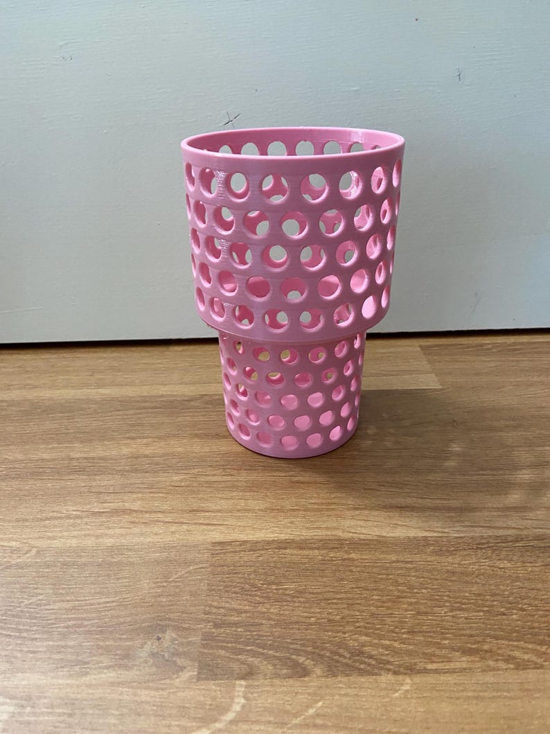 Puede incluir: Un portavasos rosa perforado con forma cil&iacute;ndrica. El portavasos tiene una serie de agujeros circulares espaciados uniformemente por todo su cuerpo. La base es ligeramente m&aacute;s peque&ntilde;a que la parte superior, creando un efecto escalonado. El art&iacute;culo es de pl&aacute;stico.