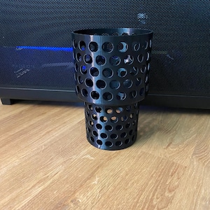 Puede incluir: Portavasos cil&iacute;ndrico negro con dise&ntilde;o perforado. El portavasos est&aacute; hecho de un material negro duro y tiene un dise&ntilde;o de dos niveles. El portavasos est&aacute; sobre una superficie de madera.