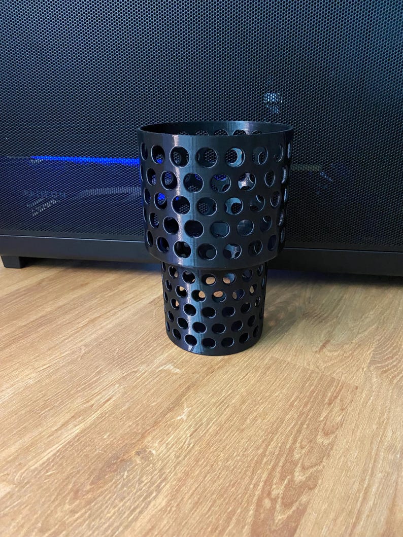Puede incluir: Portavasos cil&iacute;ndrico negro con dise&ntilde;o perforado. El portavasos est&aacute; hecho de un material negro duro y tiene un dise&ntilde;o de dos niveles. El portavasos est&aacute; sobre una superficie de madera.