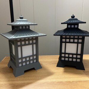 May include: Two Japanese-style lanterns, one grey and one black. Each has a pagoda-style roof and a square body with a grid pattern. The lanterns are displayed on a wooden surface.