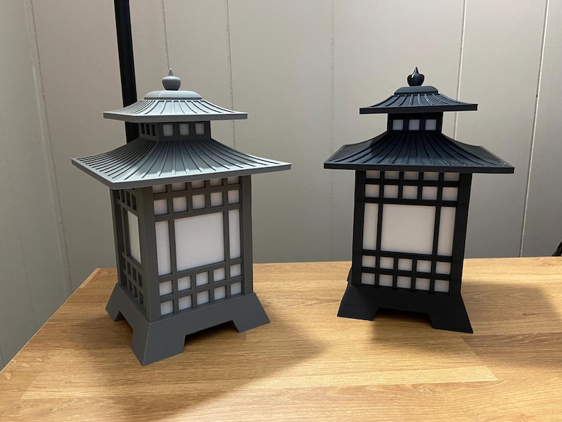 May include: Two Japanese-style lanterns, one grey and one black. Each has a pagoda-style roof and a square body with a grid pattern. The lanterns are displayed on a wooden surface.