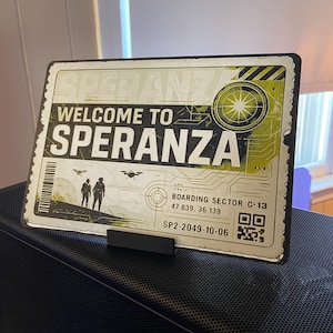 May include: A rectangular metal sign with the words "WELCOME TO SPERANZA" in large white letters. The sign has a distressed, vintage look with a green and yellow color scheme. It includes a graphic of two figures and drones.
