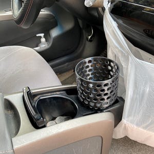 Puede incluir: Un portavasos de metal negro con un dise&ntilde;o perforado se encuentra en la consola central de un coche. La consola tambi&eacute;n cuenta con un portavasos con monedas, una palanca de cambios negra y un volante con un agarre cosido en rojo.