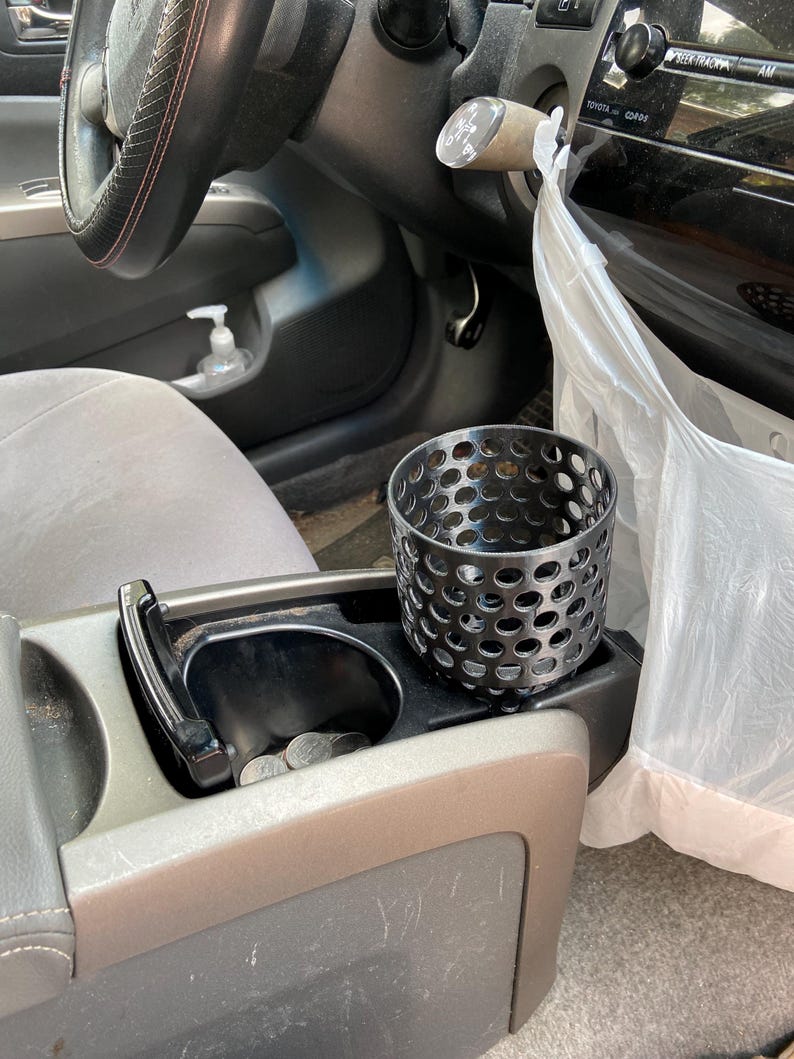 Puede incluir: Un portavasos de metal negro con un dise&ntilde;o perforado se encuentra en la consola central de un coche. La consola tambi&eacute;n cuenta con un portavasos con monedas, una palanca de cambios negra y un volante con un agarre cosido en rojo.
