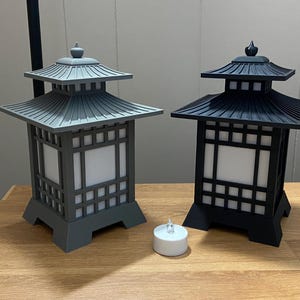 May include: Two Japanese-style lanterns, one grey and one black, with square frames and pagoda-style roofs. Each lantern has a white, illuminated centre panel. A small, white tea light candle sits in front of the lanterns.