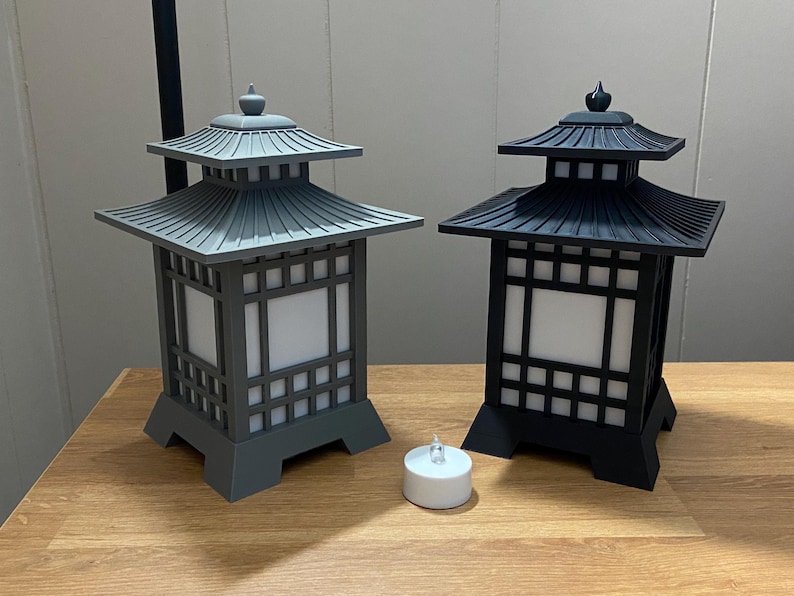 May include: Two Japanese-style lanterns, one grey and one black, with square frames and pagoda-style roofs. Each lantern has a white, illuminated centre panel. A small, white tea light candle sits in front of the lanterns.