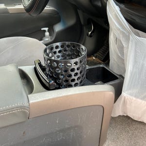 Puede incluir: Un portavasos de metal negro con un dise&ntilde;o perforado se encuentra en la consola central de un coche. El portavasos es cil&iacute;ndrico y tiene numerosos agujeros peque&ntilde;os. El interior del coche incluye un volante, asientos y una bolsa de pl&aacute;stico.